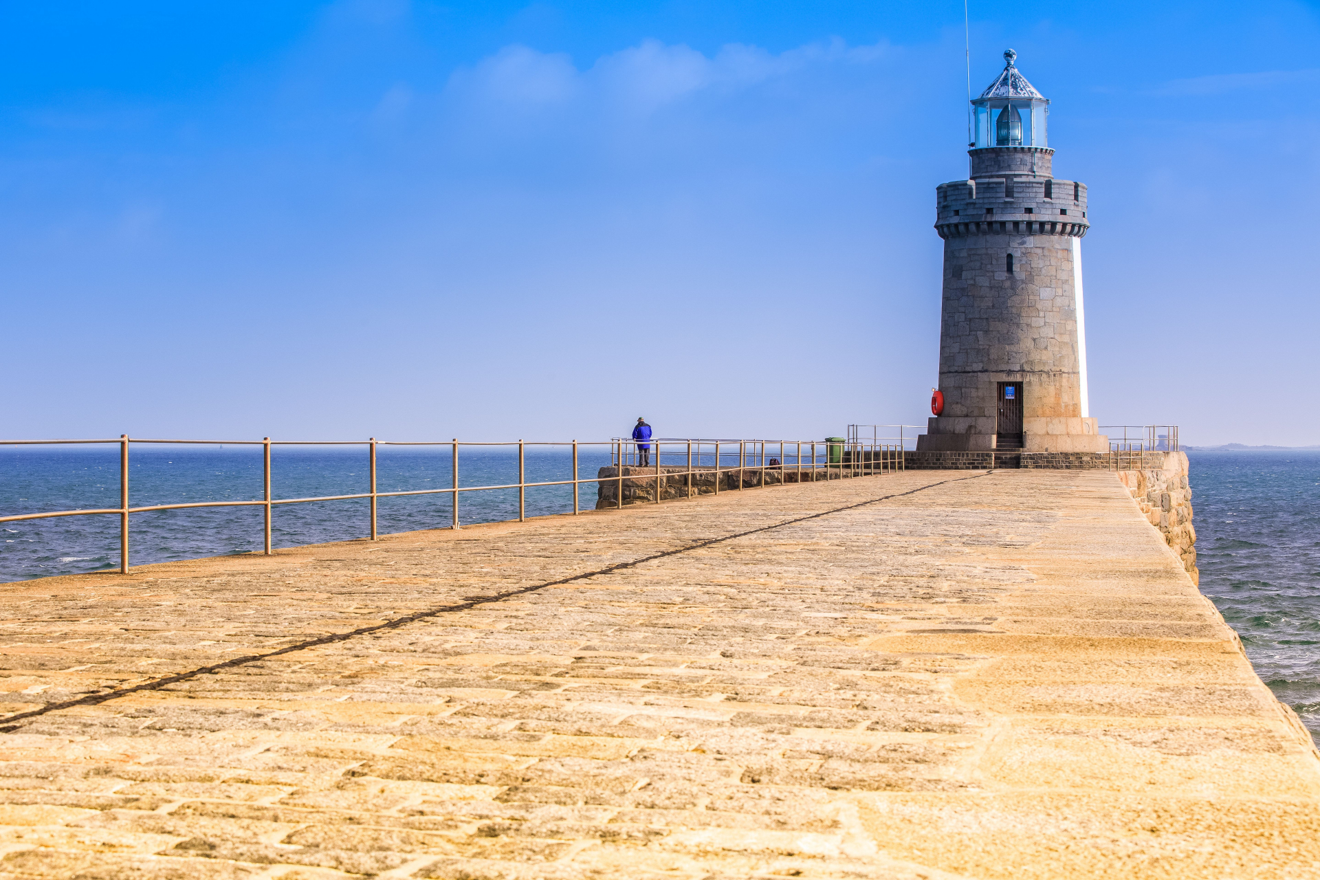 Ein breiter, sandsteinfarbener Weg führt über das Wasser zu einem Leuchtturm im Meer                                                                                            