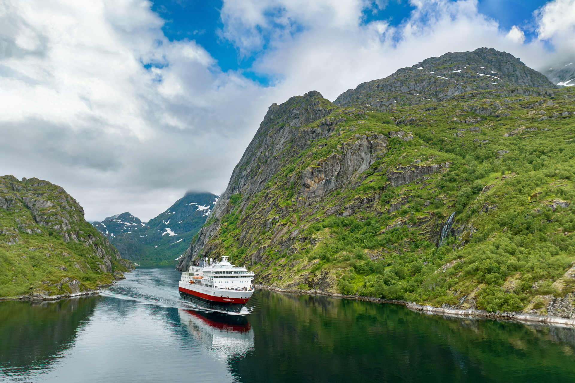 Die MS Finnmarken fährt durch einen engen, grün bewachsenen Fjord mit steilen Felswänden.                                 - © Hurtigruten | Espen Mills                                                            