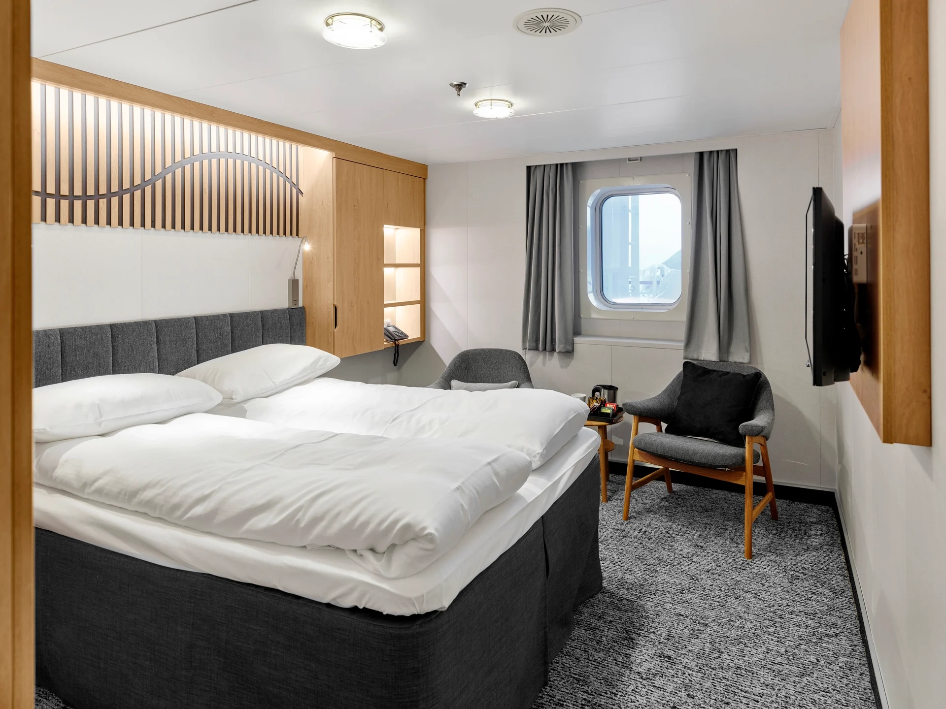 Eine helle Kabine an Bord der MS Finnmarken mit Doppelbett, grauen Polstersesseln und einem Bullauge.                                 - © Hurtigruten | Marius Viken                                                            