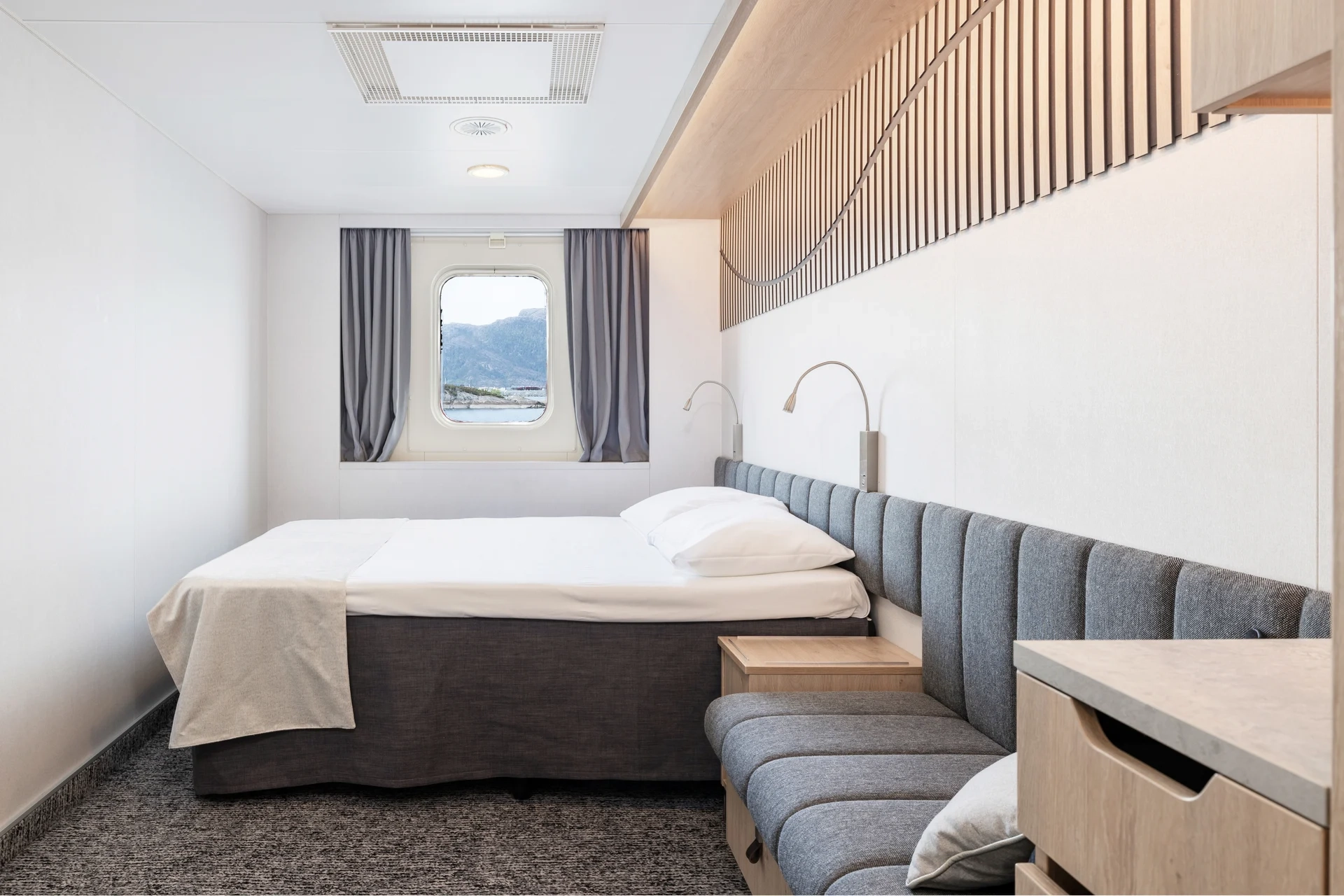 Eine helle Außenkabine an Bord der MS Finnmarken mit Doppelbett, gepolsterter Sitzbank und einem Fenster mit Bergblick.                                 - © Hurtigruten | Espen Mills                                                            